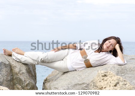 Portrait beautiful looking mature woman lying relaxed with friendly smile on rocks at ocean, with sea and blurred cloudy sky as background and copy space.