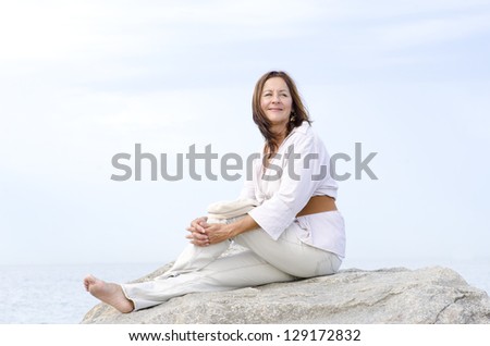 Portrait attractive mature woman, peaceful and relaxed with closed eyes, happy smiling, enjoying active retirement outdoor, isolated with ocean and white cloudy sky as background and copy space.