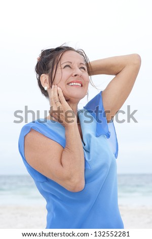 Portrait attractive mature woman relaxed and happy at beach, wearing blue blouse, with ocean and white overcast sky as blurred background and copy space.