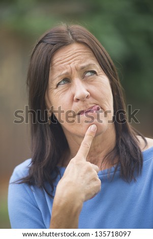 Portrait attractive mature woman outdoor with worried facial expression, thoughtful, troubled look, finger at chin and tongue out, isolated with copy space and blurred background.