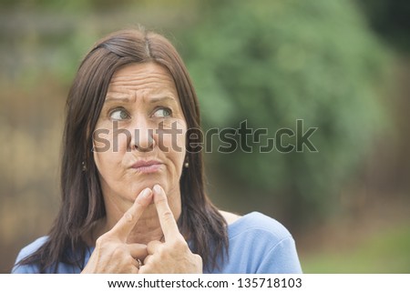 Portrait attractive mature woman outdoor with worried facial expression, thoughtful, troubled look, finger at chin, isolated with copy space and blurred background.