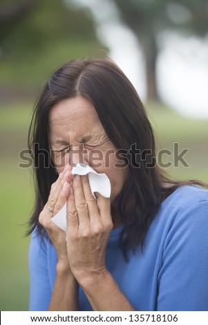 Portrait attractive mature woman suffering from cold or flu infection, sneezing into tissue, painful seasonal hayfever, with blurred outdoor background and copy space.