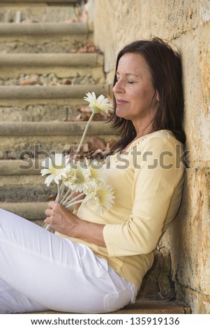Portrait beautiful mature woman with happy relaxed facial expression, in love and romance with closed eyes and flower sitting at limestone wall with blurred outdoor background.