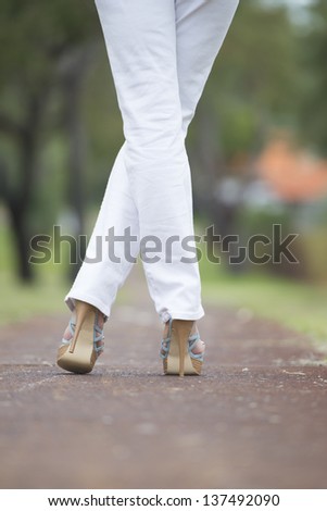 Woman with crossed legs and high heel shoes confident standing on path in park outdoor with blurred background.