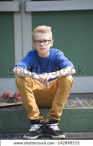 Portrait blond teenage boy with glasses sitting relaxed on steps outdoor with skateboard in background.