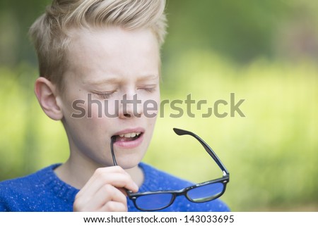 Portrait thoughtful clever and smart looking blond teenage boy with closed eyes and glasses in hand, with green blurred background outdoor.