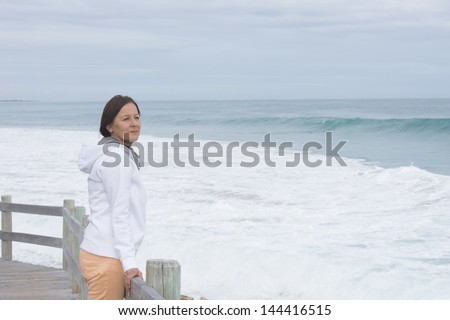 Portrait attractive mature woman standing alone at sea, looking lonely and sad, with ocean as blurred background and copy space.