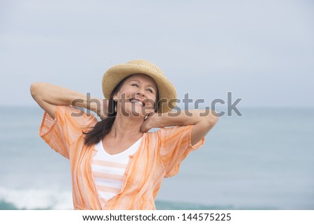 Portrait beautiful mature woman standing relaxed and joyful smiling at beach, wearing orange blouse and hat, with ocean and horizon as blurred background and copy space.