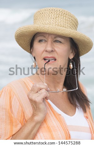 Portrait attractive mature woman with summer hat, orange blouse, thoughtful daydreaming, relaxed and confident, with blurred background outdoor.