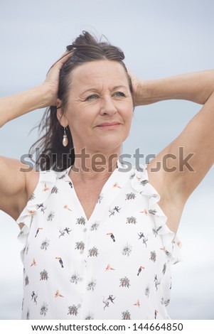 Portrait confident attractive mature woman outdoor, with arms up, hands behind neck, happy and relaxed, isolated with blurred background.
