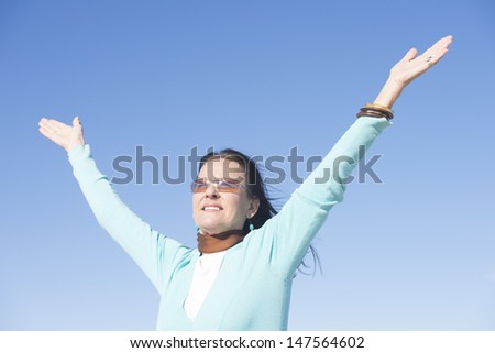 Portrait beautiful looking mature woman at sunny day outdoor, with sunglasses and hands up in air, with happy smile and confident gesture, isolated with blue sky as background and copy space.