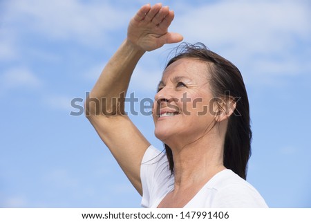 Portrait attractive mature woman with a positive outlook with a friendly and happy relaxed smile, looking up into the blue sky with hand as sunshield, with blue sky as background and copy space.