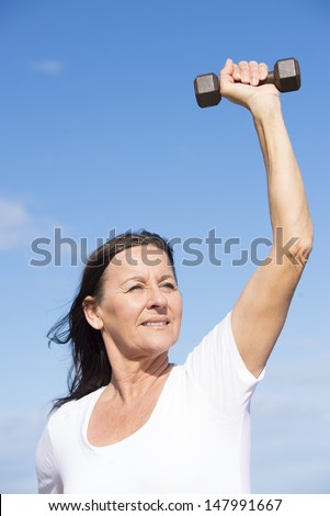 Portrait attractive confident middle aged woman exercising with weights, keeping relaxed healthy and fit, focused view, determined, positive, successful, with blue sky as background and copy space.
