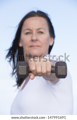 Portrait of fit and attractive mature woman in blurred background with stretched out arm with weight in focus, confident and determined, active and healthy lifestyle, with blue sky as copy space.