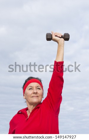 Portrait attractive mature woman showing active retirement, exercising with weights arms up outdoor, positive, confident, energetic, focused, with cloudy sky as background and copy space.