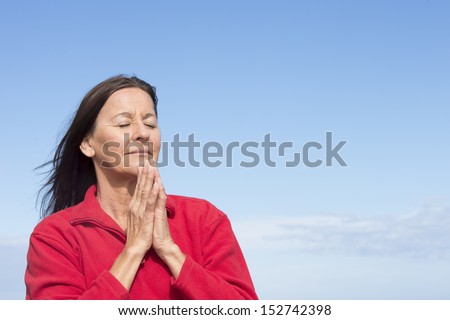 Portrait attractive mature woman relaxed, concentrated, closed eyes, smiling friendly, praying, with folded hands at chin, outdoor with blue sky as background and copy space.