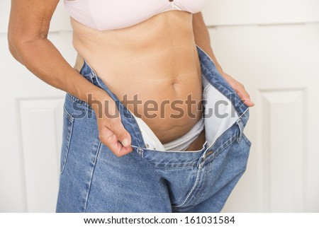Torso of mature woman showing proud result of weight loss through successful dieting, wearing over sized jeans and bra, isolated on white.
