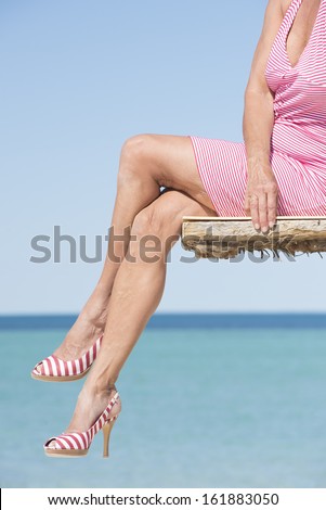 Beautiful legs of mature woman sitting in sexy pose on platform at the ocean, wearing summer dress and high heels, overlooking tropical sea, with horizon and blue sky as background and copy space.