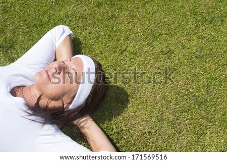 Portrait sporty mature woman lying relaxed with closed eyes and hands behind neck on a sunny summer day on green grass, with copy space.