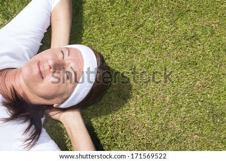 Portrait sporty and attractive senior woman lying relaxed with closed eyes and hands behind neck on a sunny summer day on green grass, with copy space.