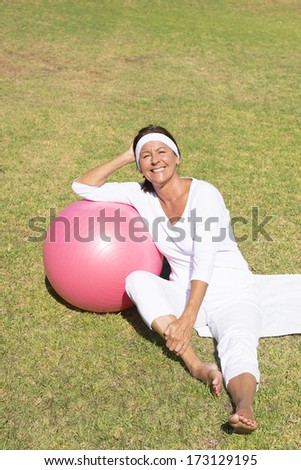 Portrait fit and active senior woman sitting happy relaxed leaning on gymnastic ball on green grass.
