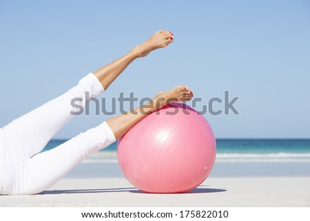 Womans legs up in the air with pink gymnastic ball, exercising at beach, with  ocean and blue sky as background and copy space.
