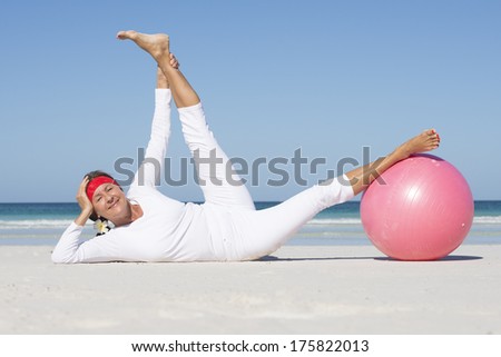 Portrait active and fit attractive looking mature woman at beach doing stretch exercises with gymnastic ball, legs up in the air, ocean and blue sky as blurred background and copy space.