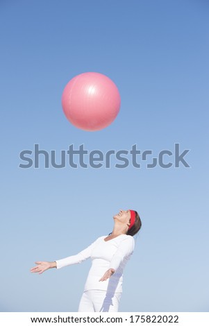 Happy, healthy and sporty attractive mature woman catching gymnastic ball while exercising, with blue sky as background and copy space.