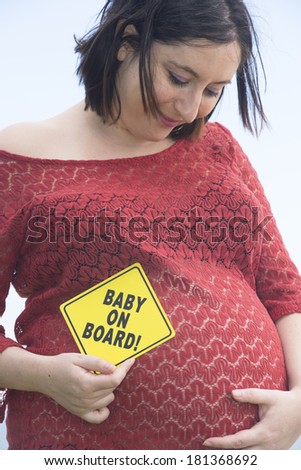 Portrait pregnant woman holding \'baby on board\' sign at belly, blurred background and copy space.