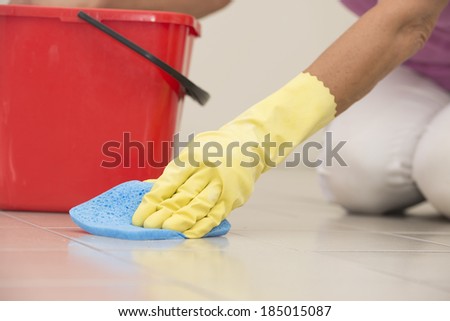 Close up Hand in yellow rubber glove cleaning floor tiles with sponge, with woman, housewife in blurred background and copy space.