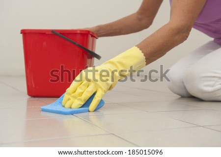 Hand in yellow rubber glove cleaning floor tiles with sponge, with woman, housewife in blurred background and copy space.
