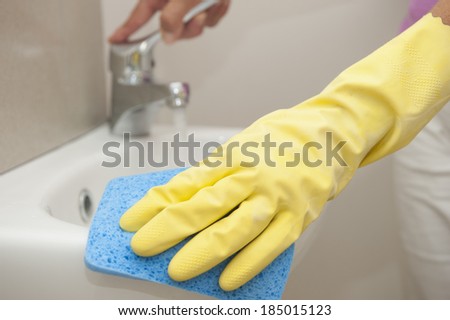 Hand in yellow rubber glove cleaning bathroom basin with blue sponge, with faucet or tap in blurred background and with copy space.
