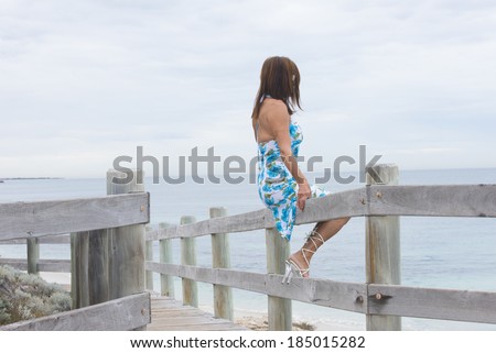 Portrait attractive mature woman sitting lonely at beach overlooking thoughtful ocean in blurred background, wearing summer dress and high heel shoes
