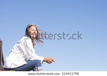 Portrait friendly smiling attractive mature woman, joyful sitting on bench at sunny windy day outdoor, with blue sky as background and copy space.