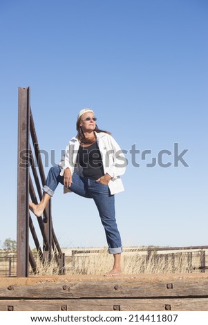 Portrait of attractive mature woman, standing confident and laid back bare feet outdoor, wearing jeans, jacket, sunglasses and hat, with horizon and blue sky as copy space and background.