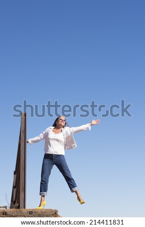 Portrait attractive mature woman, confident, joyful, happy, with arm up, leg up, wearing high heel shoes outdoor, with blue sky as background and copy space.