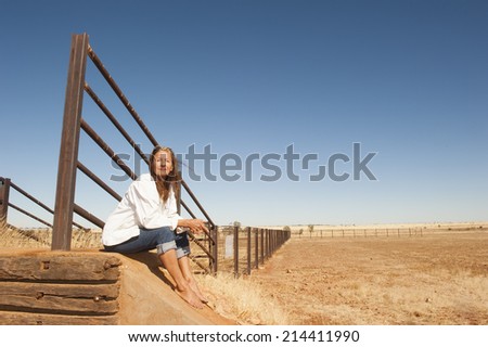 Portrait attractive mature woman sitting relaxed at metal fence line in rural farming area in outback Australia, with dry arid agricultural country and blue sky as background and copy space.