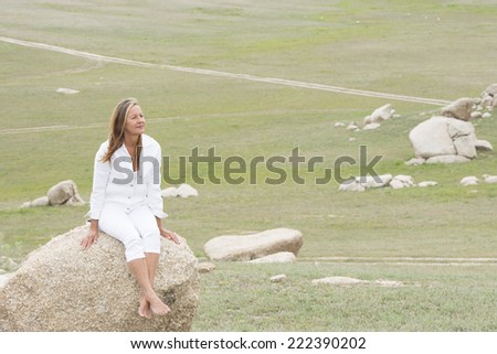 Portrait of attractive mature woman sitting happy and relaxed bare feet in remote countryside landscape on rock, wearing white clothes, with grass field  as background and copy space.