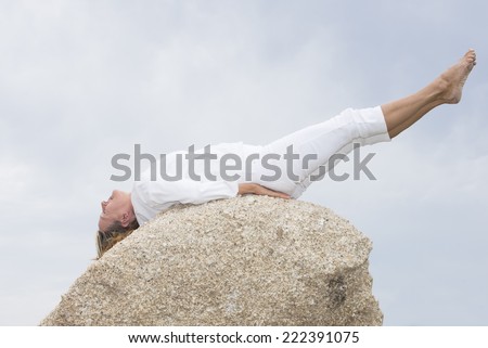 Portrait attractive retired mature woman doing stretching yoga or pilates exercises outdoor on rock, with legs up, body stretched, concentrated, closed eyes, copy space.