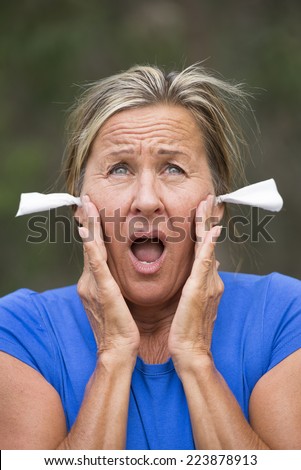 Portrait attractive mature woman with stressed suffering facial expression, with tissues in ears for noise protection, outdoor blurred background.