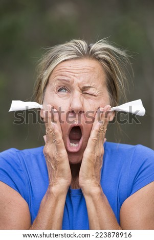 Portrait attractive mature woman with stressed shocked facial expression, with tissues as earplugs for noise protection, outdoor blurred background.