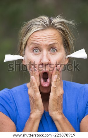 Portrait attractive mature woman with stressed shocked facial expression, with tissues in ears for noise protection, outdoor blurred background.