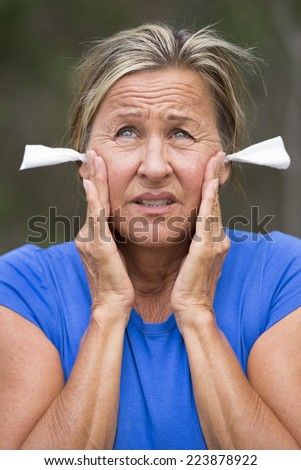 Portrait attractive mature woman with stressed facial expression, with tissues in ears for noise protection, outdoor blurred background.