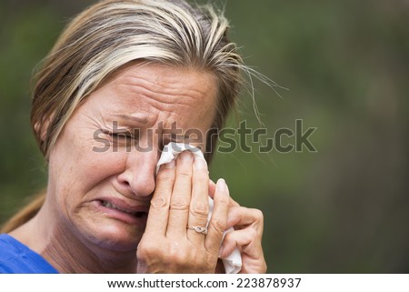Portrait crying mature woman in grief, with sad unhappy, stressed emotional facial expression, suffering painful depression, tissue in hands, outdoor blurred background.
