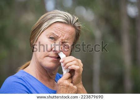 Portrait attractive mature woman with tissue stuffed in running nose, suffering  from cold, flu or hay fever allergy, one eye closed, outdoor blurred background.
