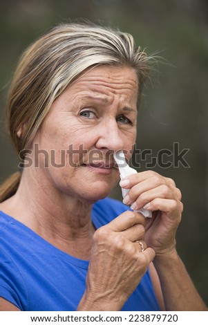 Portrait attractive mature woman with tissue stuffed in running nose, suffering  unhappy from cold, flu or hay fever allergy, outdoor blurred background.