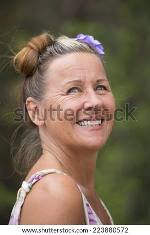 Portrait of attractive mature woman with flower in hair, confident smiling, relaxed and joyful with green blurred background outdoor.