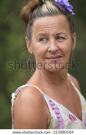 Portrait of attractive mature woman with flower in hair, confident smiling, relaxed and laid back with green blurred background outdoor.