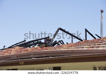 Fire damaged roof on house building, with charred black timber beams, broken tiles and chimney, blue sky as background and copy space.