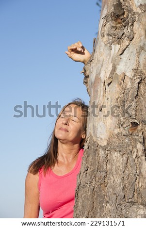 Portrait happy relaxed attractive mature woman leaning against tree outdoor with arm up and closed eyes, blue sky as background and copy space.